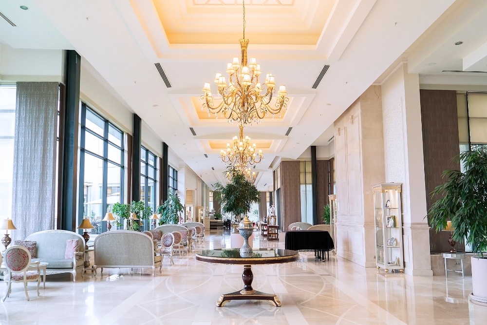 A long hallway with a crystal chandelier hanging from a high ceiling. The hallway has cream colored walls and dark wood furniture, including chairs, tables, and a couch. This image is relevant to a search for the best commercial paint because it depicts a luxurious commercial lobby, a common area that benefits from a high-quality paint job. The image also shows neutral-colored walls, a popular choice for commercial interiors.