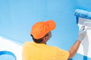 Man in an orange cap and yellow shirt painting a fibreglass pool with a blue roller, demonstrating the practical steps in how to paint a fibreglass pool for an even and professional finish.