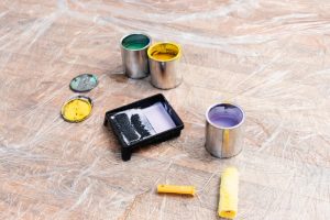 Paint cans with leftover green, yellow, and purple paint, along with a paint tray and roller on a plastic-covered floor, illustrating creative leftover paint ideas for home decor projects