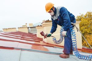 Worker in protective gear using a spray gun to apply paint on a corrugated metal roof, demonstrating preventive measures against roof crack paint issues.