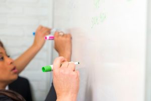 Close-up of two people writing with markers on a whiteboard wall painted with writable paint for walls, emphasizing the practical use of writable surfaces in office environments.