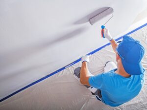 A worker in a blue cap and gloves using a roller to apply paint to a wall, demonstrating wall painting techniques.