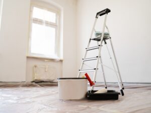 Room prepared for painting a house with ladder, paint bucket, and tools placed on protective plastic covering the floor.