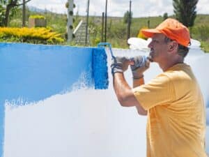 A man repainting a fibreglass pool with a roller, applying a fresh blue coat to the pool wall.