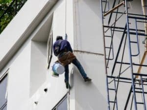 Worker hanging on rope repairing flaking paint on tall building exterior wall.