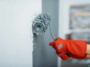 Professional applying grey coat during new home painting using a roller brush and red gloves on a white wall.