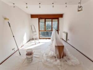 A room prepped to paint a room, with drop sheets, paint roller, buckets, and a ladder present.