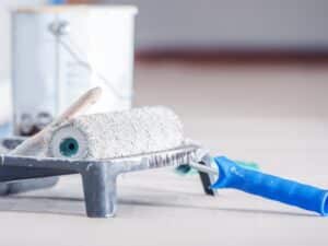 A paint roller and tray, ready to paint a room, sit on the floor near a paint can.