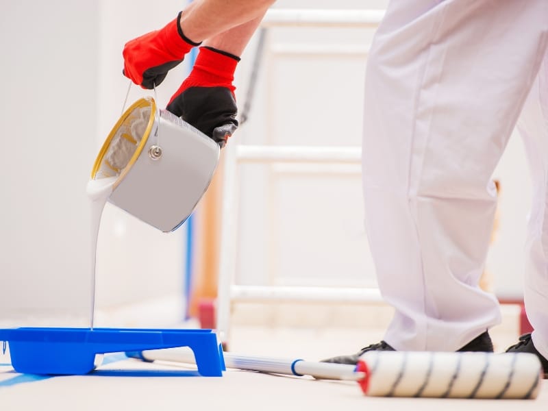 Close-up of a painter wearing gloves pouring white paint into a tray, illustrating the preparation stage of kitchen painting.